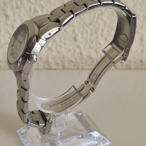 May include: A silver stainless steel wristwatch with a round face and a linked band. The watch is displayed on a clear acrylic stand. The words "STAINLESS STEEL" are visible on the band.