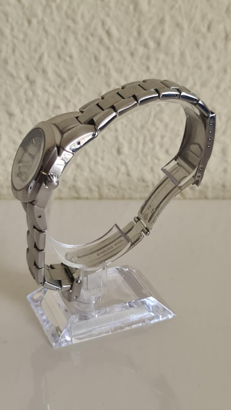 May include: A silver stainless steel wristwatch with a round face and a linked band. The watch is displayed on a clear acrylic stand. The words "STAINLESS STEEL" are visible on the band.