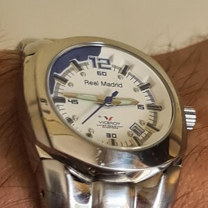 May include: A silver-toned wristwatch with a white face and blue accents. The watch features the text "Real Madrid" and the brand name "Viceroy". The watch has a stainless steel band and a round face.