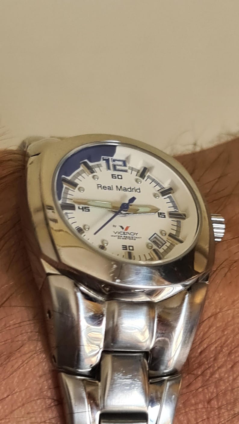 May include: A silver-toned wristwatch with a white face and blue accents. The watch features the text "Real Madrid" and the brand name "Viceroy". The watch has a stainless steel band and a round face.