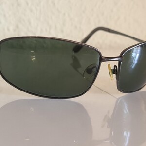 May include: Sunglasses with a dark metallic frame and rectangular lenses in a deep green tint. The eyewear features thin metal temples and adjustable gold-toned nose pads, providing a structured look with a reflective finish on the lenses.