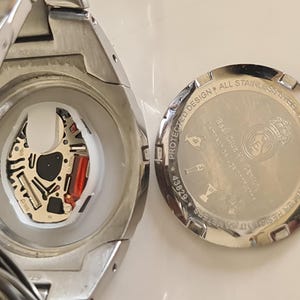 May include: Close-up of a silver wristwatch with the back cover removed, revealing the internal mechanism. The watch face is visible, along with the intricate circuitry and a red component. The back cover is also visible, with text and a logo.