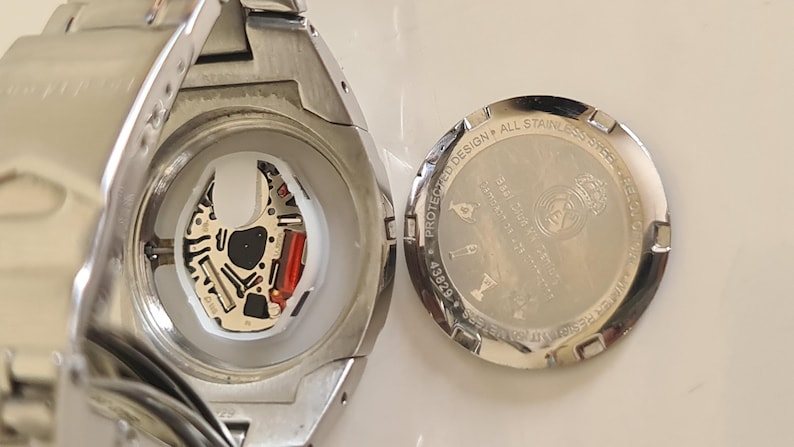 May include: Close-up of a silver wristwatch with the back cover removed, revealing the internal mechanism. The watch face is visible, along with the intricate circuitry and a red component. The back cover is also visible, with text and a logo.