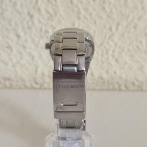 May include: A silver-toned wristwatch with a metal band and clasp. The watch is displayed on a clear, square stand. The clasp is engraved with the text "Real Madrid".