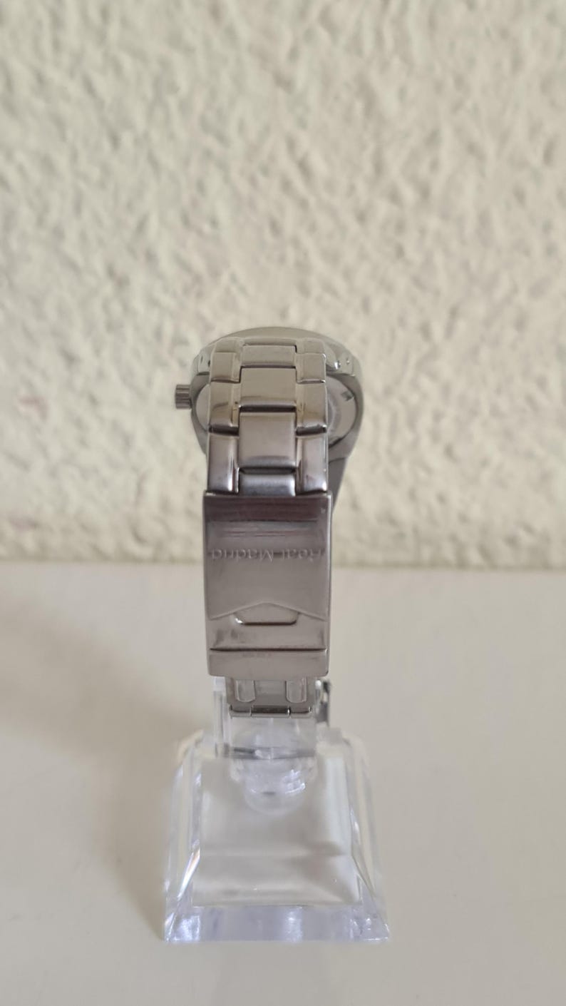 May include: A silver-toned wristwatch with a metal band and clasp. The watch is displayed on a clear, square stand. The clasp is engraved with the text "Real Madrid".