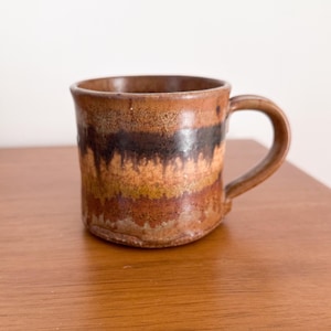 May include: A handmade ceramic mug with a brown, black, and orange striped design. The mug has a rounded base and a curved handle. The mug is sitting on a wooden surface.