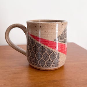 May include: A speckled beige ceramic mug featuring a curved handle and a decorative pattern of black leaf-like shapes arranged in a grid. A bold, diagonal red stripe cuts across the center of the mug, contrasting with the textured clay surface.