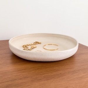 May include: A shallow, round, off-white ceramic trinket dish with a smooth, matte finish. Inside the dish rests a gold-toned metal chain necklace with a small circular pendant and a thin, open gold-toned metal bangle bracelet.