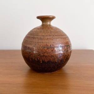 May include: A small, spherical ceramic vase with a narrow neck and flared rim. The surface features a two-tone glaze, transitioning from a light tan, textured upper section with horizontal ridges to a dark, mottled brown base.