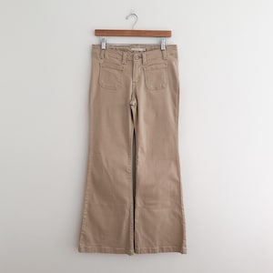 May include: Tan-colored flared pants with two front pockets and a button closure, hanging on a wooden hanger. The pants have a classic, casual style, suitable for everyday wear. The fabric appears to be a sturdy cotton blend.