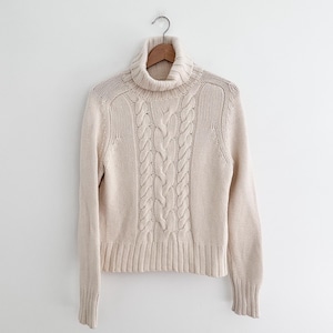 May include: Cream-colored cable knit turtleneck sweater hanging on a wooden hanger. The sweater features a ribbed hem and cuffs, with a prominent cable knit design down the front. The neutral color and classic design make it a versatile wardrobe staple.