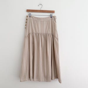 May include: A beige midi skirt with vertical stripes and a gathered waist. The skirt hangs from a wooden hanger and features decorative buttons on the side. The skirt is made of a lightweight fabric.