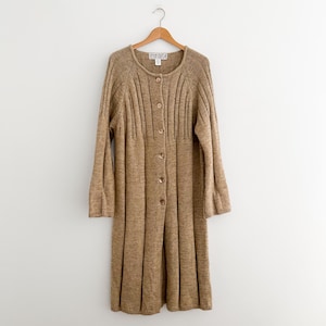 May include: A long-sleeved, knee-length tan cardigan sweater featuring a round neckline, a vertical ribbed knit pattern on the upper bodice, and a button-down front with six matching buttons. The garment is displayed on a wooden clothes hanger.