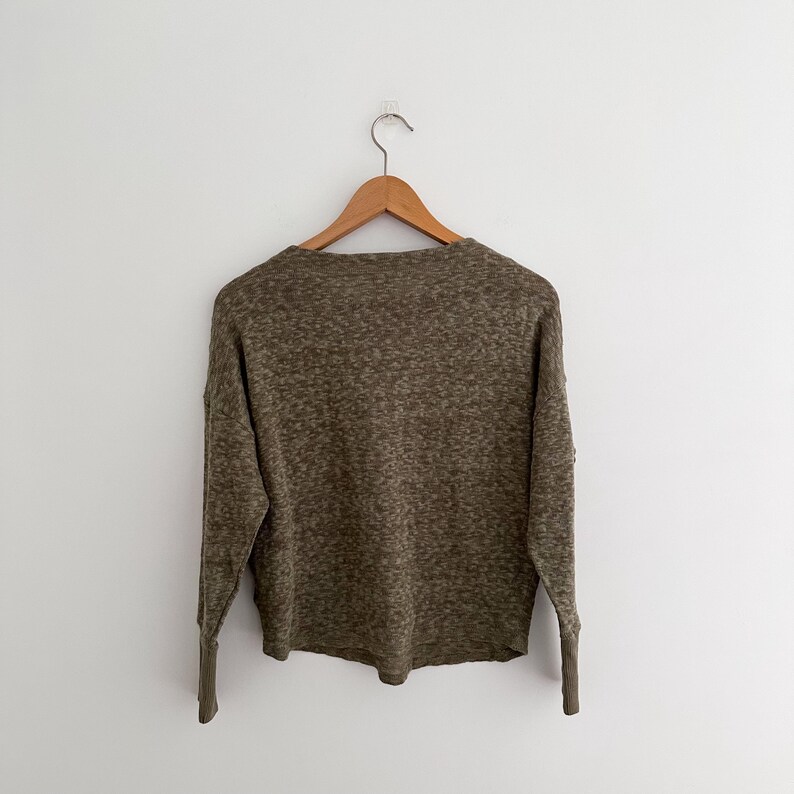 May include: A long-sleeved olive green sweater with a speckled pattern, hanging on a wooden hanger against a white wall. The sweater has a crew neck and ribbed cuffs. The overall style is casual and comfortable.