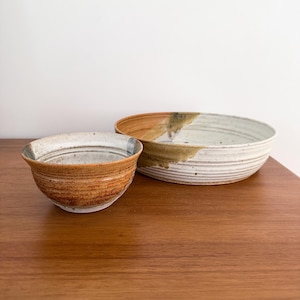 May include: Two handmade ceramic bowls. The smaller bowl is a warm brown with a textured surface and a lighter interior. The larger bowl is a cream color with brown and green accents and a wide, shallow shape. Both bowls are on a wooden surface.