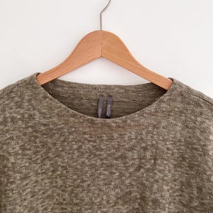 May include: Olive green knit top with a crew neck, hanging on a wooden hanger. The fabric has a textured appearance. The top is a neutral color, suitable for various outfits.