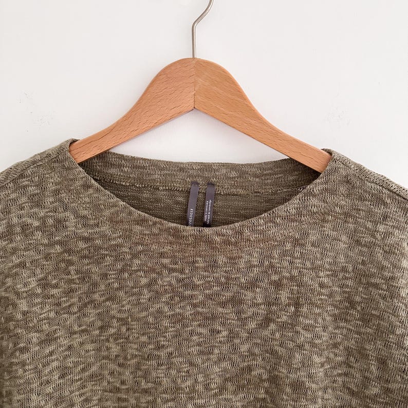 May include: Olive green knit top with a crew neck, hanging on a wooden hanger. The fabric has a textured appearance. The top is a neutral color, suitable for various outfits.