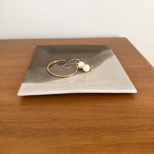 May include: A square ceramic tray featuring a two-tone design with a dark charcoal gray section and a light gray section. Resting on the tray are several pieces of gold-toned jewelry, including a thin bangle, two small rings, and two pearl earrings.