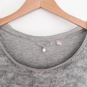 May include: Close-up of a gray knit top with a round neckline and a sheer, light gray trim. A small, silver pendant hangs from the neckline. The top is hanging on a wooden hanger against a white background.