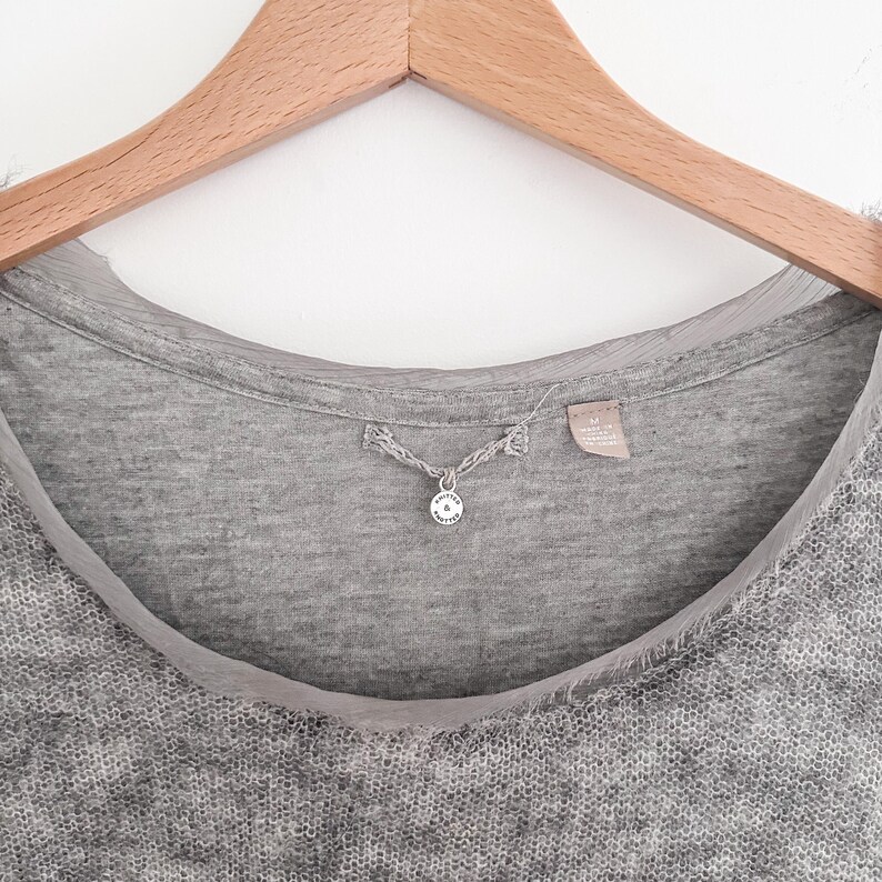 May include: Close-up of a gray knit top with a round neckline and a sheer, light gray trim. A small, silver pendant hangs from the neckline. The top is hanging on a wooden hanger against a white background.