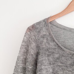 May include: A light gray, long-sleeved sweater with a sheer, open knit texture. The sweater has a scoop neck and is hanging on a wooden hanger against a white background. The neckline is trimmed with a lighter gray fabric.