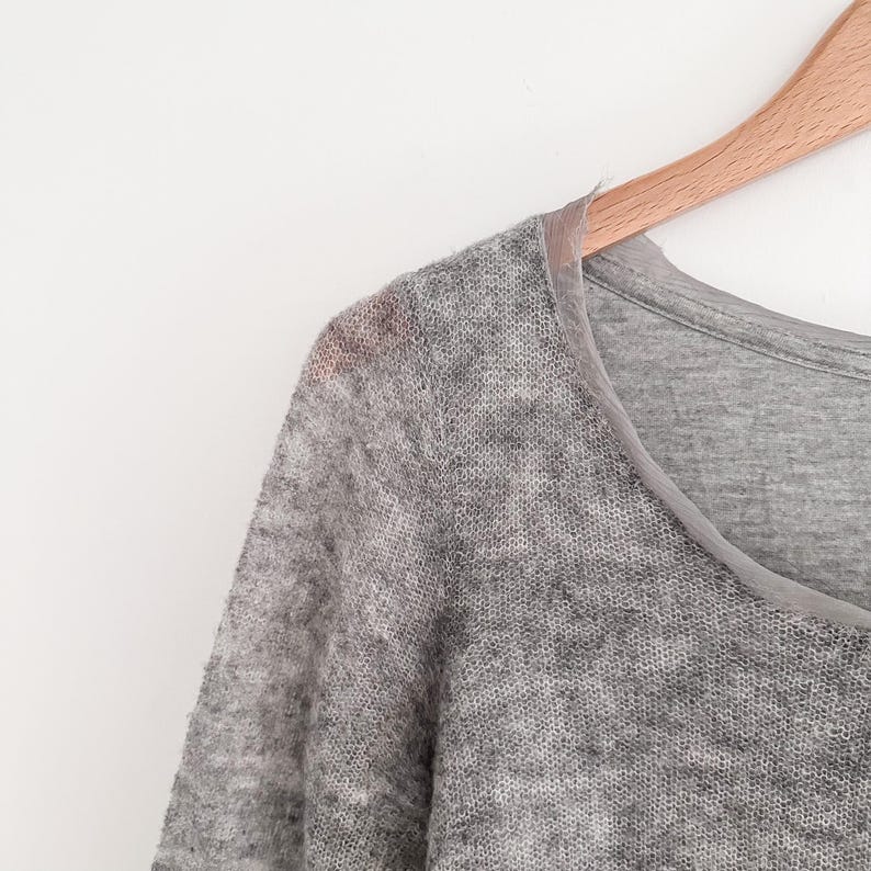 May include: A light gray, long-sleeved sweater with a sheer, open knit texture. The sweater has a scoop neck and is hanging on a wooden hanger against a white background. The neckline is trimmed with a lighter gray fabric.