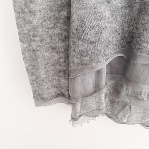 May include: Close-up of a light gray knit fabric with a layered, sheer, fringed trim. The fabric has a soft, textured appearance. The layers of fabric create a flowing, draped effect. The overall aesthetic is delicate and airy.