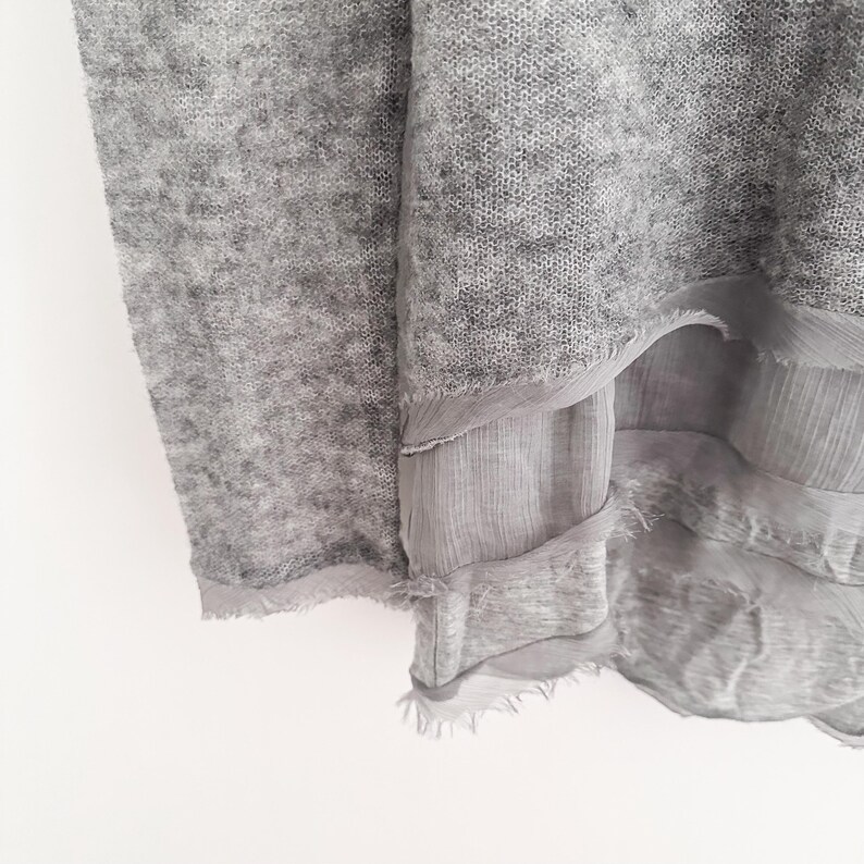 May include: Close-up of a light gray knit fabric with a layered, sheer, fringed trim. The fabric has a soft, textured appearance. The layers of fabric create a flowing, draped effect. The overall aesthetic is delicate and airy.