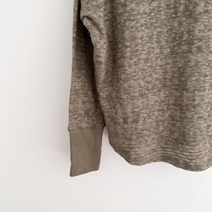 May include: Close-up of a long-sleeved, olive green sweater. The fabric has a textured, woven appearance. The cuff is a solid olive green, ribbed knit. The sweater is a neutral color, suitable for various outfits.