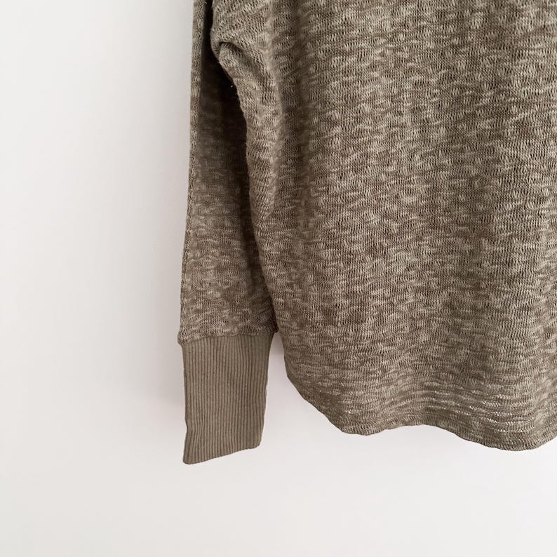 May include: Close-up of a long-sleeved, olive green sweater. The fabric has a textured, woven appearance. The cuff is a solid olive green, ribbed knit. The sweater is a neutral color, suitable for various outfits.