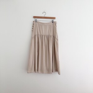 May include: A beige and white striped skirt hangs on a wooden hanger. The skirt has a high waist, a gathered detail at the waistline, and a flared hem. Gold-colored buttons adorn the side of the skirt.