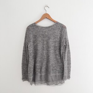 May include: A gray sweater with a round neckline and long sleeves hangs on a wooden hanger. The sweater has a layered design, with a sheer, ruffled hem. The fabric appears soft and textured, suitable for cooler weather.