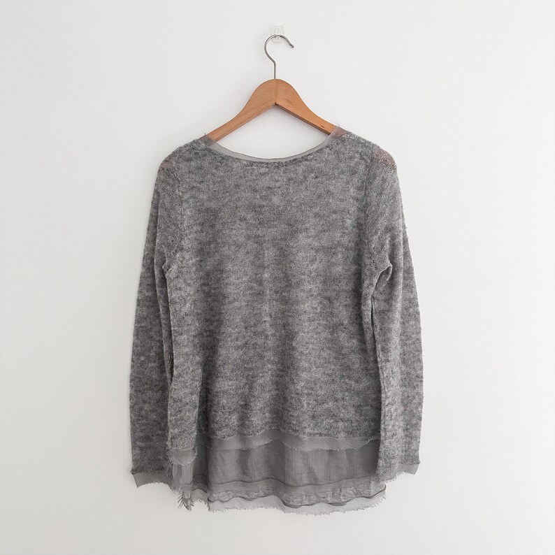 May include: A gray sweater with a round neckline and long sleeves hangs on a wooden hanger. The sweater has a layered design, with a sheer, ruffled hem. The fabric appears soft and textured, suitable for cooler weather.