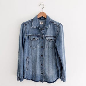 May include: Light blue denim jacket with long sleeves, two chest pockets, and two side pockets. The jacket has a button-down front and a frayed hem. The jacket is hanging on a wooden hanger.