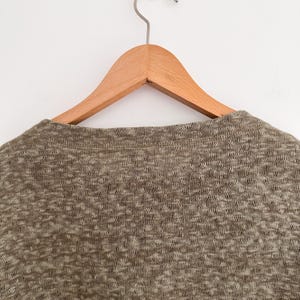 May include: Close-up of a heathered knit top in shades of brown and beige, hanging on a wooden hanger. The fabric has a textured appearance. The hanger is suspended from a metal hook against a white background.