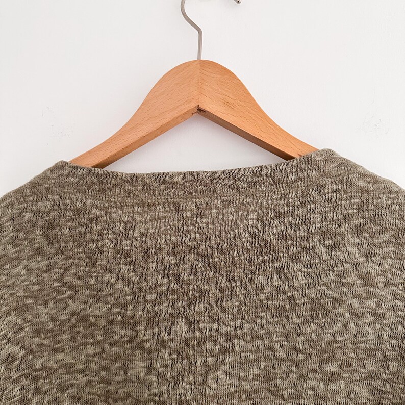 May include: Close-up of a heathered knit top in shades of brown and beige, hanging on a wooden hanger. The fabric has a textured appearance. The hanger is suspended from a metal hook against a white background.