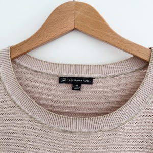 May include: Close-up of a light pink knit sweater with a scoop neck and a ribbed collar. The sweater is hanging on a wooden hanger. The label reads "ADRIANNA PAPELL".