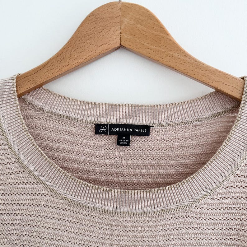 May include: Close-up of a light pink knit sweater with a scoop neck and a ribbed collar. The sweater is hanging on a wooden hanger. The label reads "ADRIANNA PAPELL".