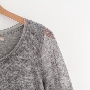 May include: A gray, long-sleeved sweater with a V-neck design. The sweater has a textured, open-knit pattern and a contrasting fabric detail on the shoulder. The garment is hanging on a wooden hanger.