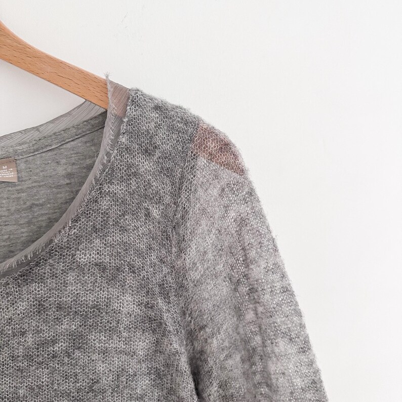 May include: A gray, long-sleeved sweater with a V-neck design. The sweater has a textured, open-knit pattern and a contrasting fabric detail on the shoulder. The garment is hanging on a wooden hanger.