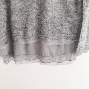 May include: A light gray sweater with a layered, sheer, gray fabric detail at the bottom. The sweater has a textured appearance, and the sheer fabric has a frayed edge. The background is white.