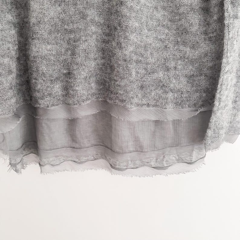 May include: A light gray sweater with a layered, sheer, gray fabric detail at the bottom. The sweater has a textured appearance, and the sheer fabric has a frayed edge. The background is white.
