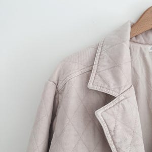 May include: A light beige quilted jacket with a classic collar and diamond stitching. The jacket is hanging on a wooden hanger. The neutral color and quilted design give the jacket a timeless appeal.