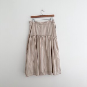 May include: A beige and white striped skirt hangs on a wooden hanger. The skirt has a high waist and a flared, midi-length design. The fabric has vertical stripes in a neutral color palette.