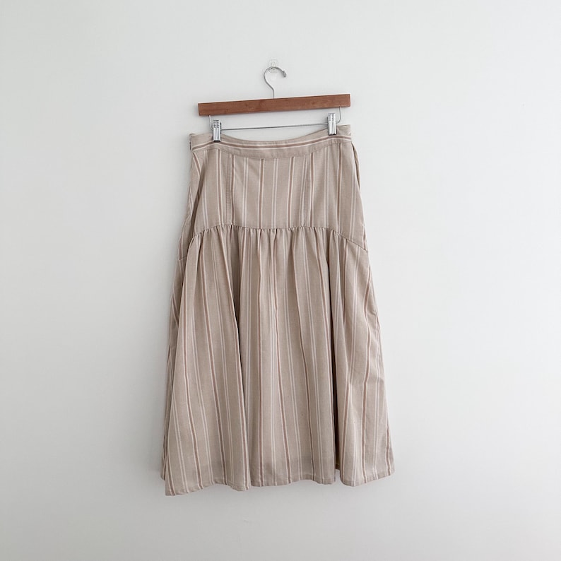 May include: A beige and white striped skirt hangs on a wooden hanger. The skirt has a high waist and a flared, midi-length design. The fabric has vertical stripes in a neutral color palette.