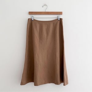 May include: A brown midi skirt hanging on a wooden hanger. The skirt has a slight A-line shape and a seam down the front. The fabric appears to be a smooth, solid color. The skirt is hanging against a white background.