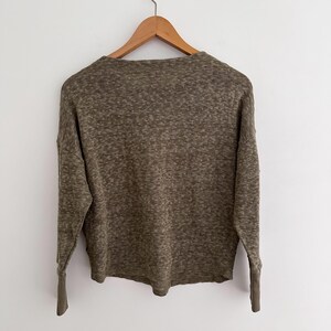 May include: Olive green long-sleeve sweater with a textured knit design. The sweater is hanging on a wooden hanger against a white background. The sleeves have ribbed cuffs.