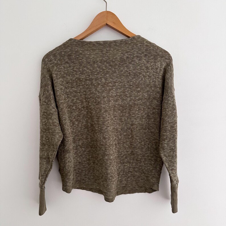 May include: Olive green long-sleeve sweater with a textured knit design. The sweater is hanging on a wooden hanger against a white background. The sleeves have ribbed cuffs.