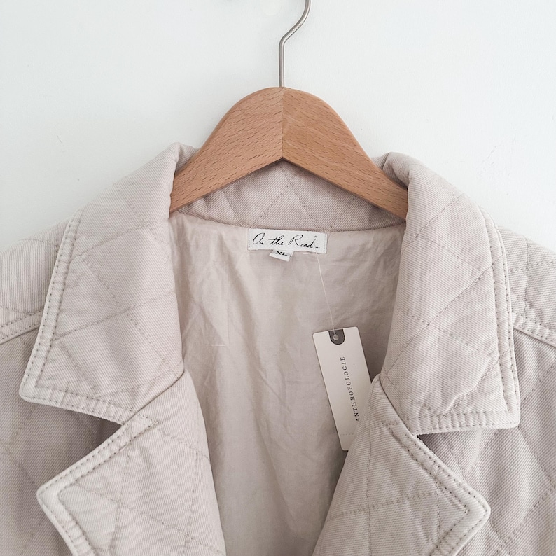 May include: A light beige quilted jacket with a diamond pattern, hanging on a wooden hanger. The jacket has a collar and a tag that reads "On the Road" and "Anthropologie".
