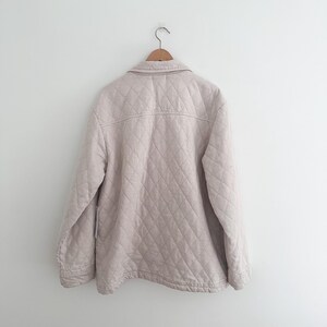 May include: A light beige quilted jacket hanging on a wooden hanger. The jacket has a diamond pattern and a button-down front. The jacket is made of a soft, lightweight material, perfect for layering.